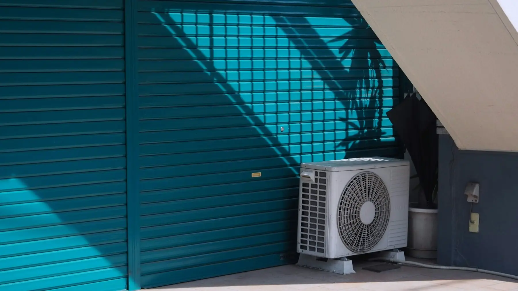 Outdoor air conditioning unit on its feet beside a closed metal shutter in bright sun