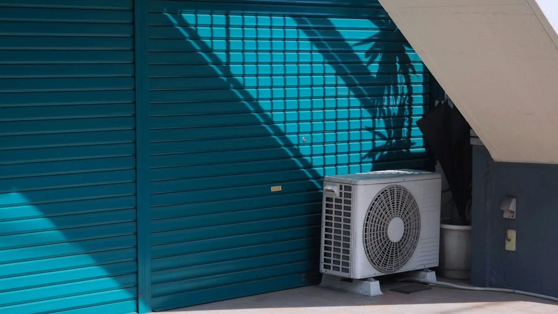 Outdoor air conditioning unit on its feet beside a closed metal shutter in bright sun