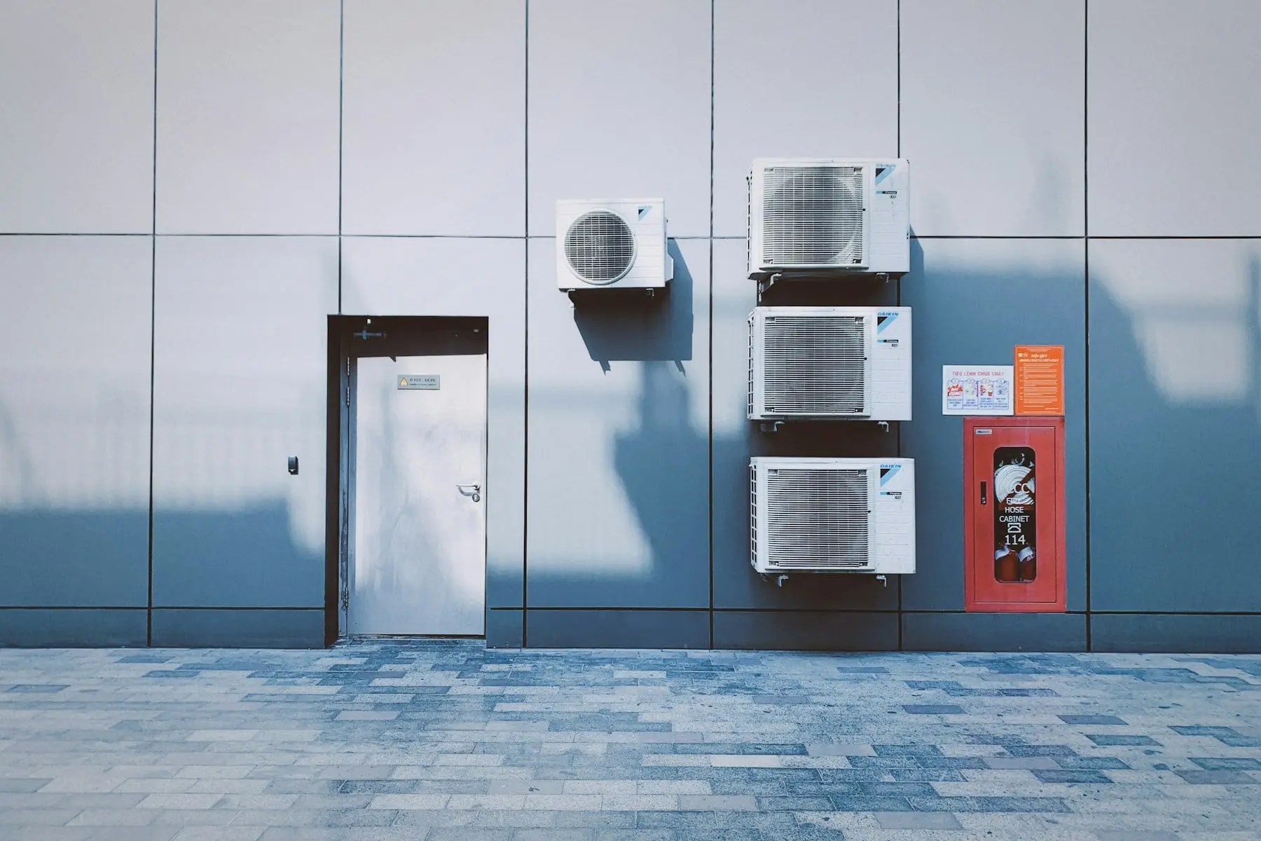 Four outdoor air conditioning units mounted on a grey panelled wall beside a door