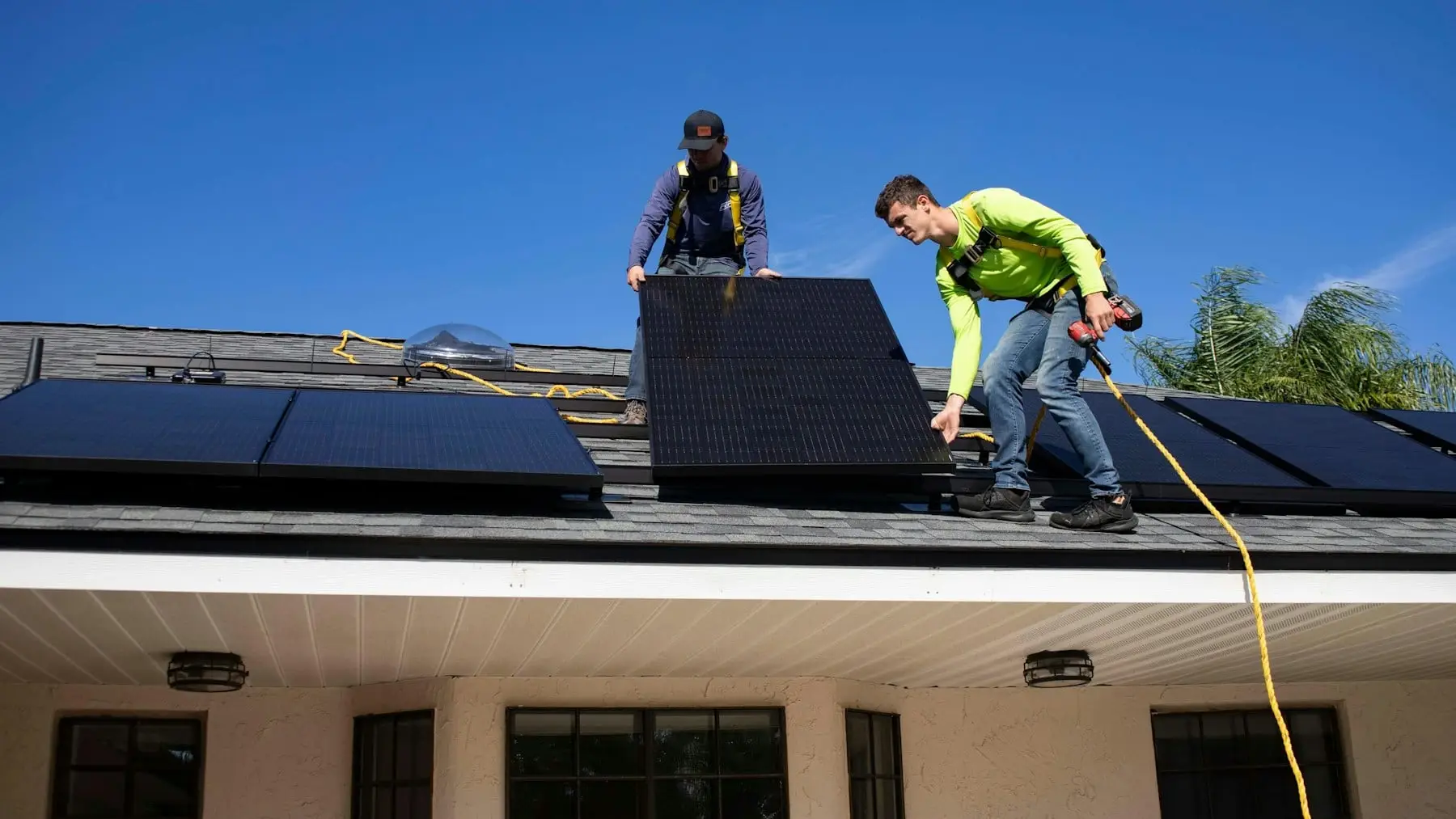 Two installers in safety harnesses fitting black solar panels on a house roof